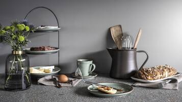 A kitchen counter with a tea set, a plate of pastries, a vase with flowers, and a metal container with utensils.