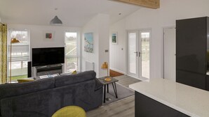 A modern living room with a sofa, TV, and a dining area with a table. - 11 Meadow Retreat (Liskeard)