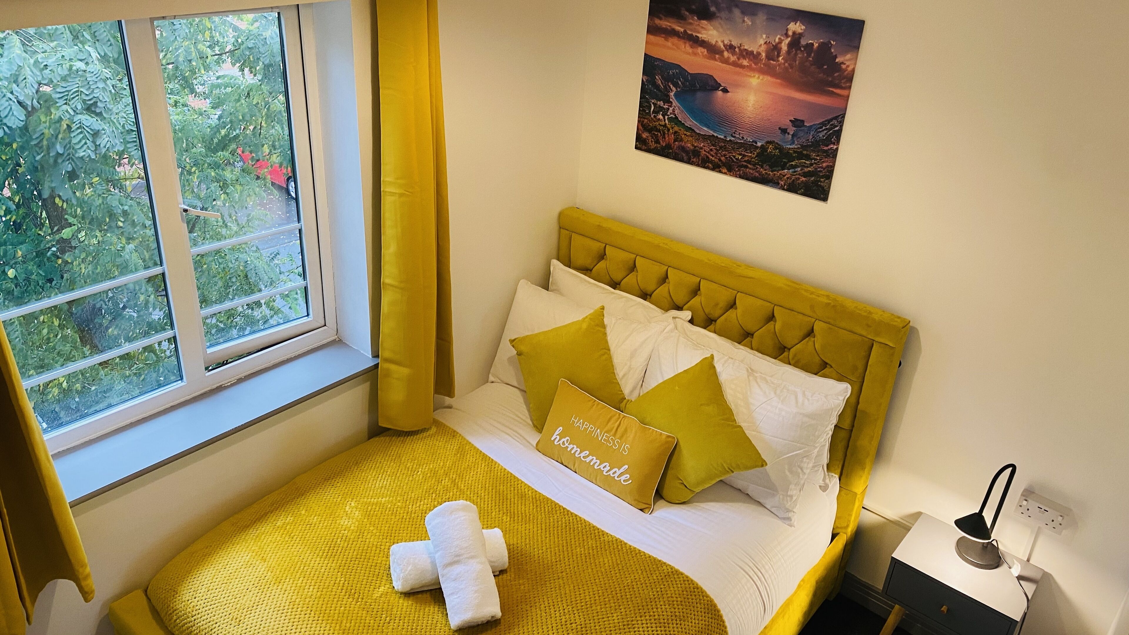 Un dormitorio con cama, ventana, un cuadro y mesita de noche.