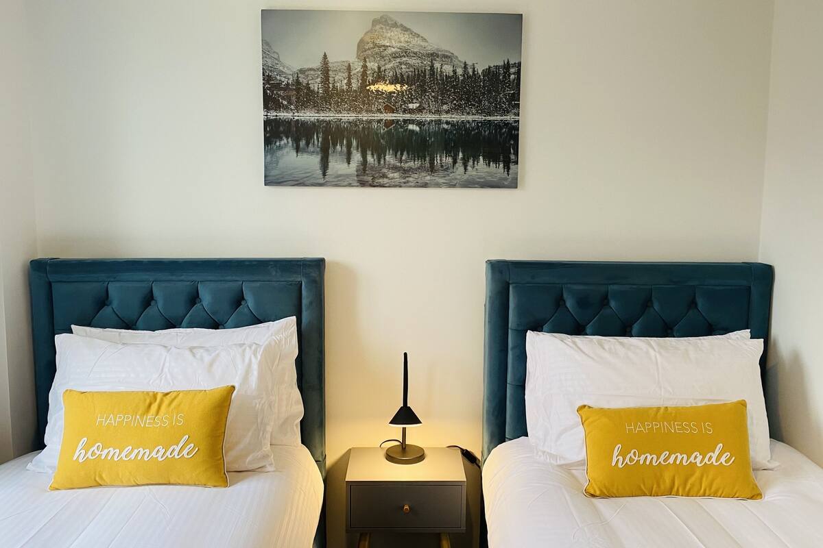 A hotel room with two beds, each with a blue headboard and white bedding. There are two yellow pillows with the text "HAPPINESS IS homemade." A small bedside table with a lamp is between the beds. Above the beds, there is a framed picture of a snowy mountain landscape.