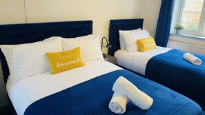 A hotel room with two beds, each with blue bedding and white pillows. One bed has a yellow pillow with the text "Happiness is homemade." There are towels on the beds and a window with a view of greenery outside. - Cosy Stay In Birmingham by Villazu (Birmingham)