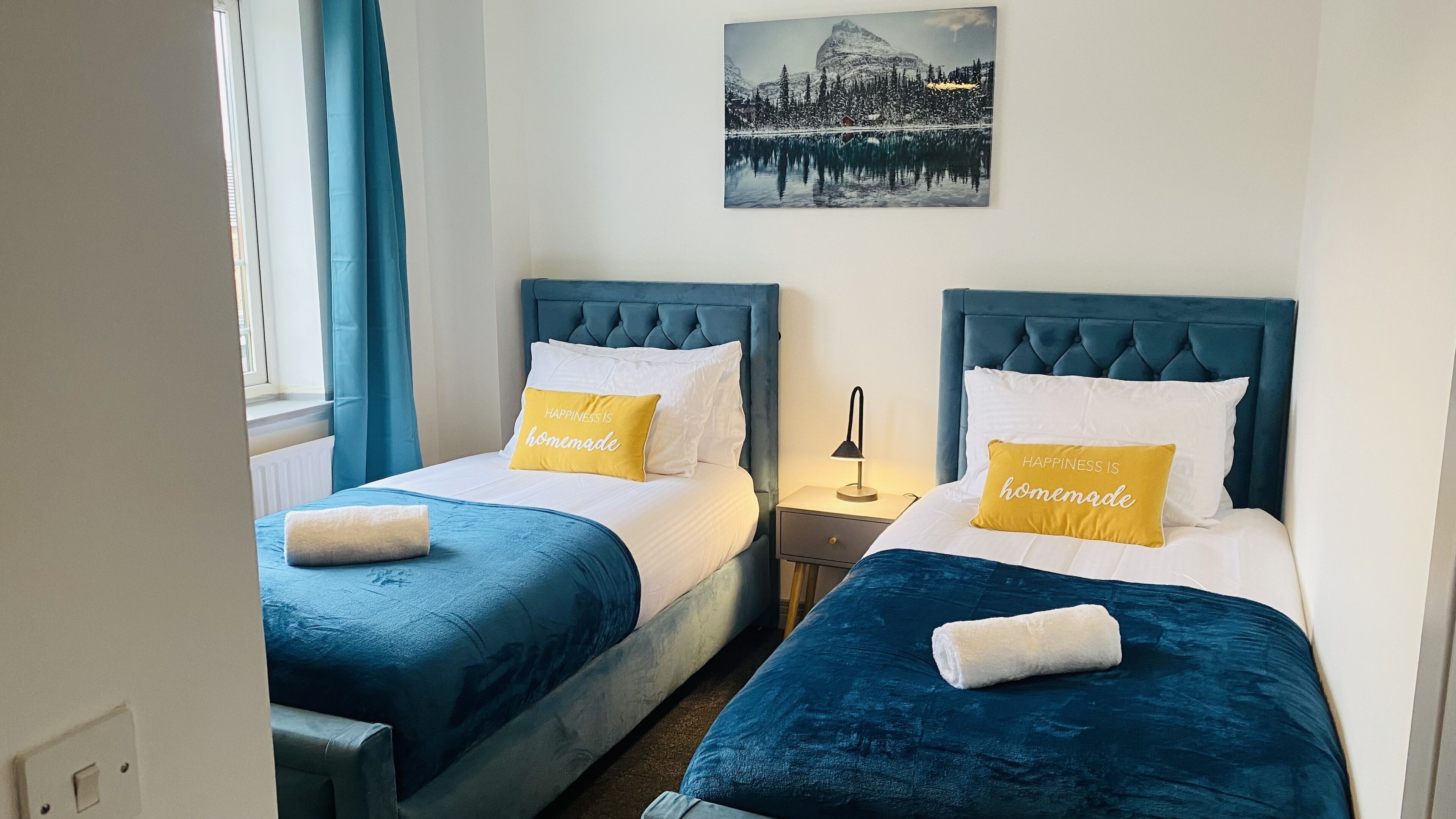 A hotel room with two beds, each with a blue headboard and white bedding. There are two yellow pillows with the text "HAPPINESS IS homemade" on them. A painting of a snowy landscape is visible on the wall above the beds.
