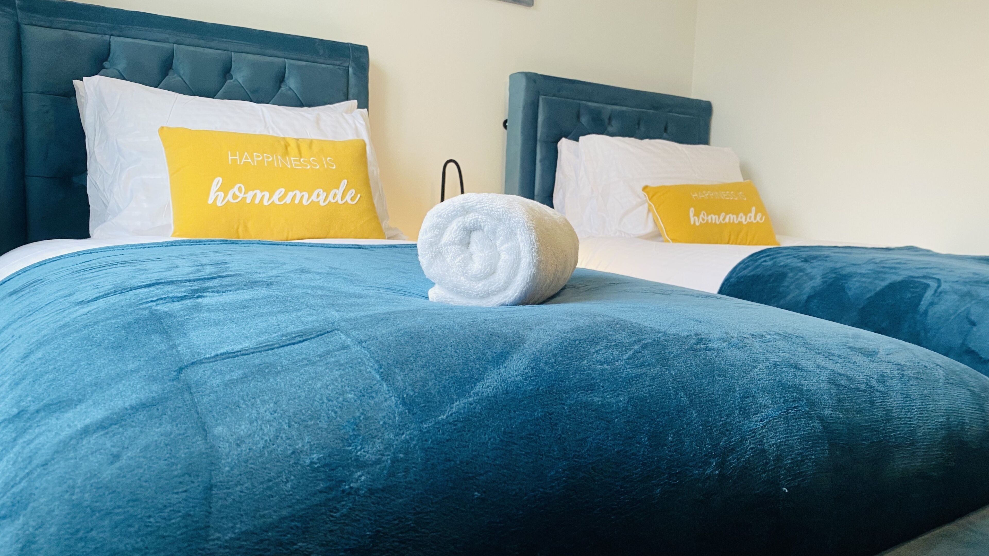 Une chambre d’hôtel avec deux lits, chacun doté d’une tête de lit bleue et d’un oreiller orné de la phrase suivante : « Le bonheur, ça se crée soi-même ».