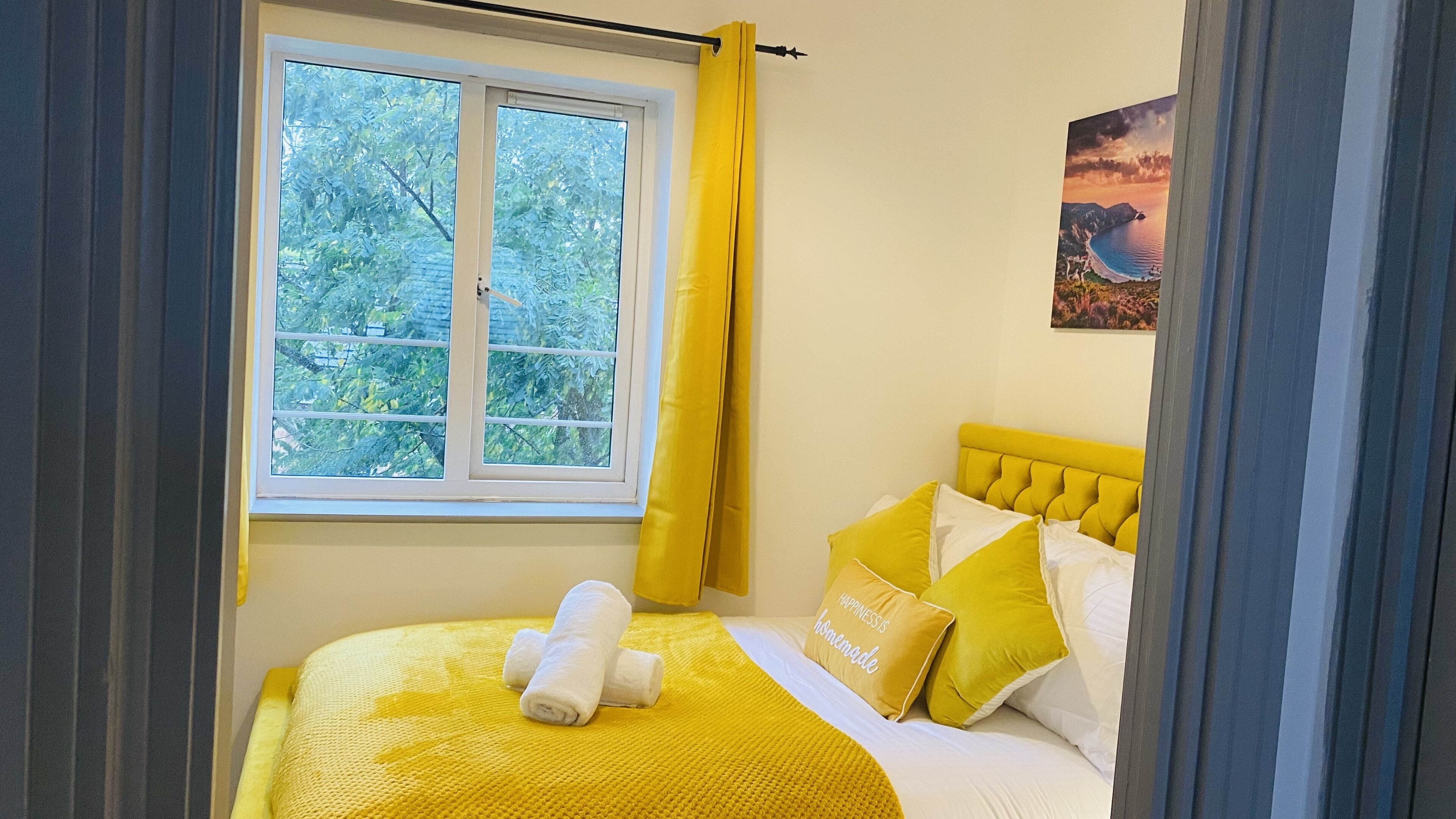 A bedroom with a bed, yellow curtains, a window with a view of trees, and a painting on the wall.
