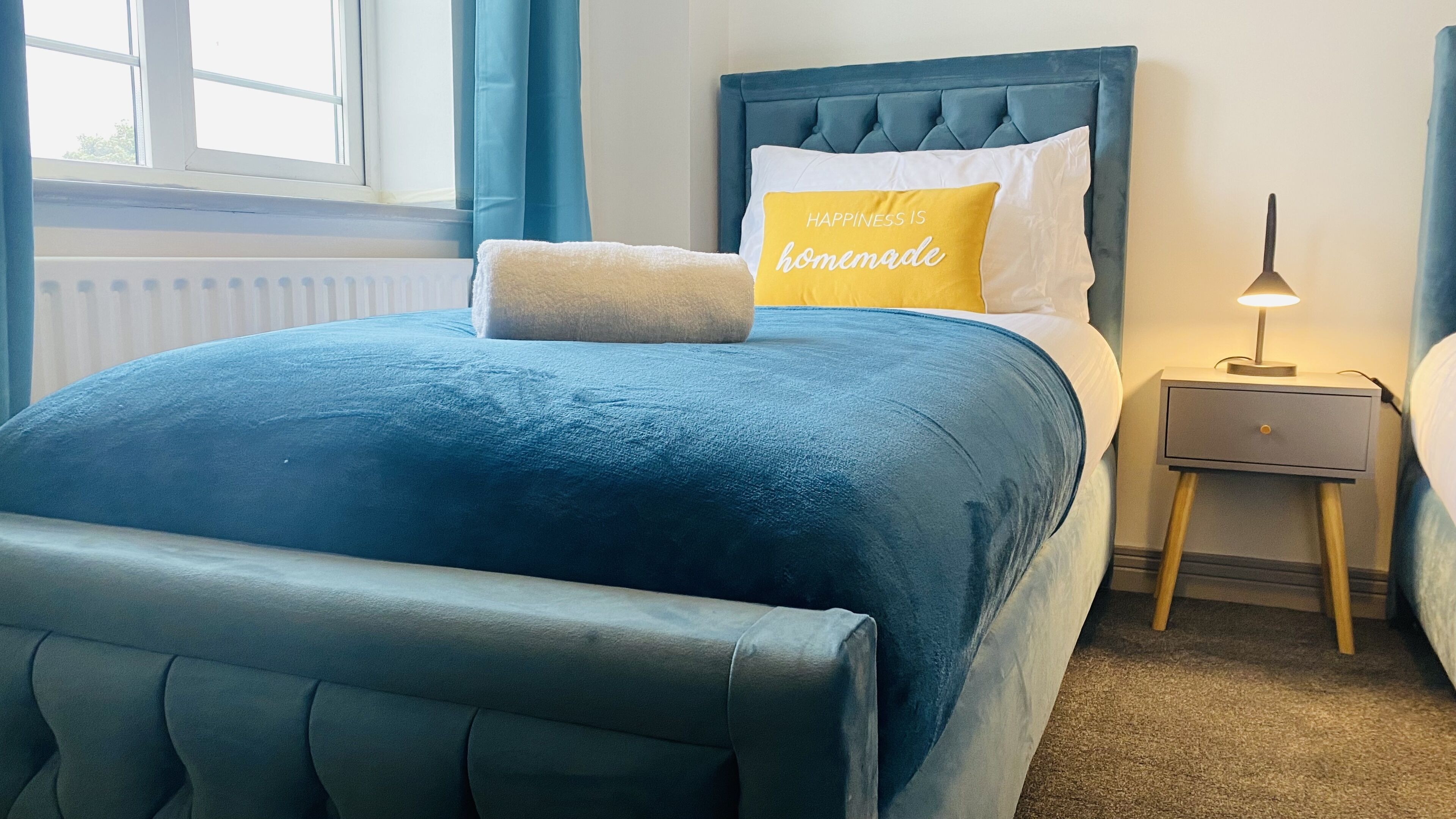 Cama bem arrumada com edredom azul, travesseiro branco e travesseiro amarelo com a inscrição 'Happiness is homemade'.  Tem uma mesa de cabeceira com uma luminária e uma gavetinha.
