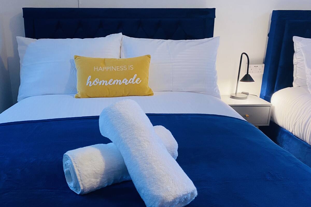 A neatly made bed with a blue bedspread, a white pillow with 'HAPPINESS IS homemade' text, and a rolled white towel.