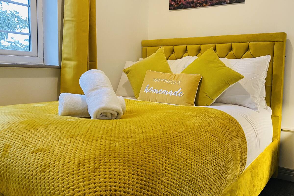 A bed with a yellow headboard, a rolled white towel, and pillows with a 'happiness is homemade' message.