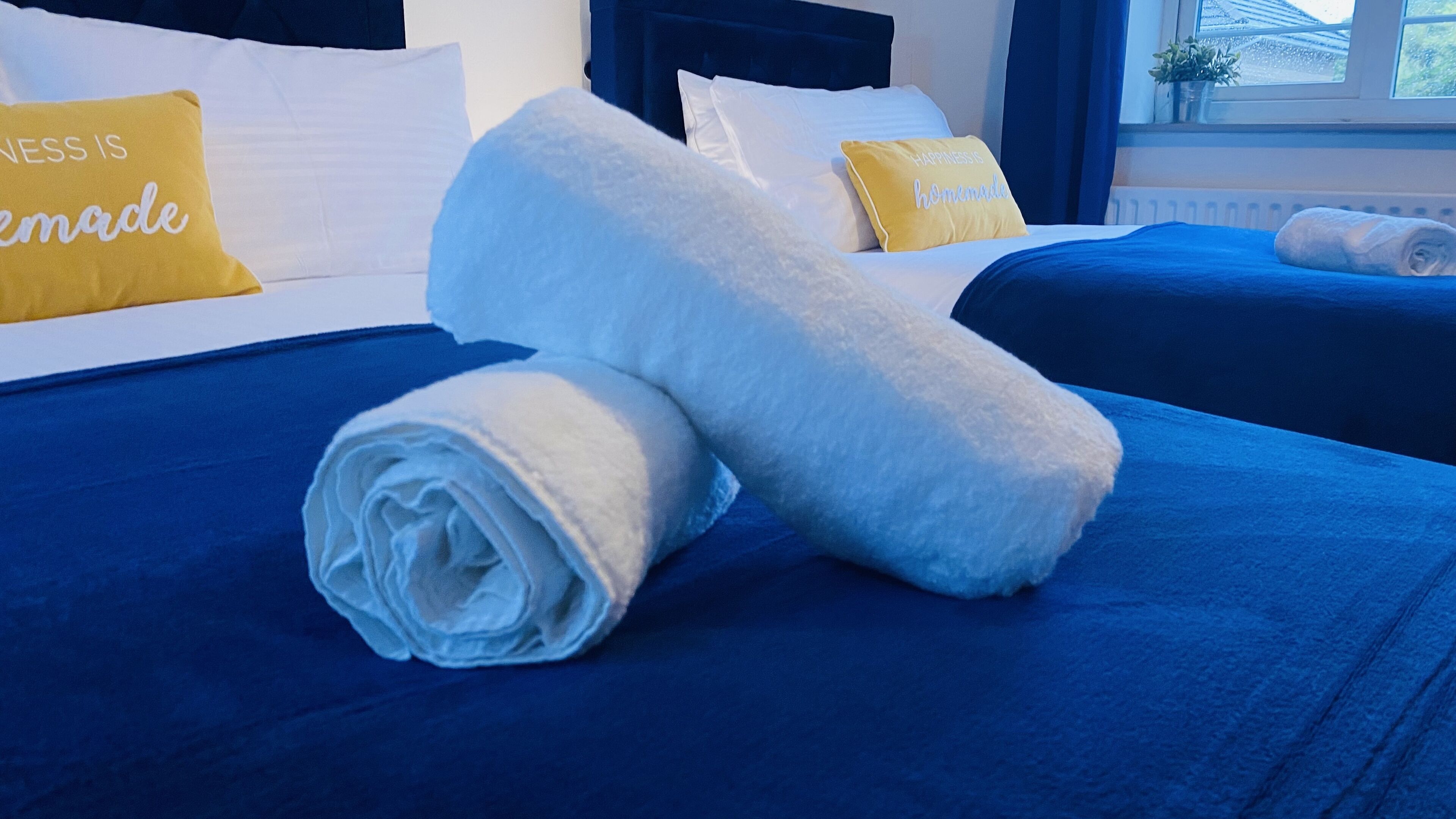 Una cama bien tendida con ropa de cama azul y dos toallas blancas enrolladas.