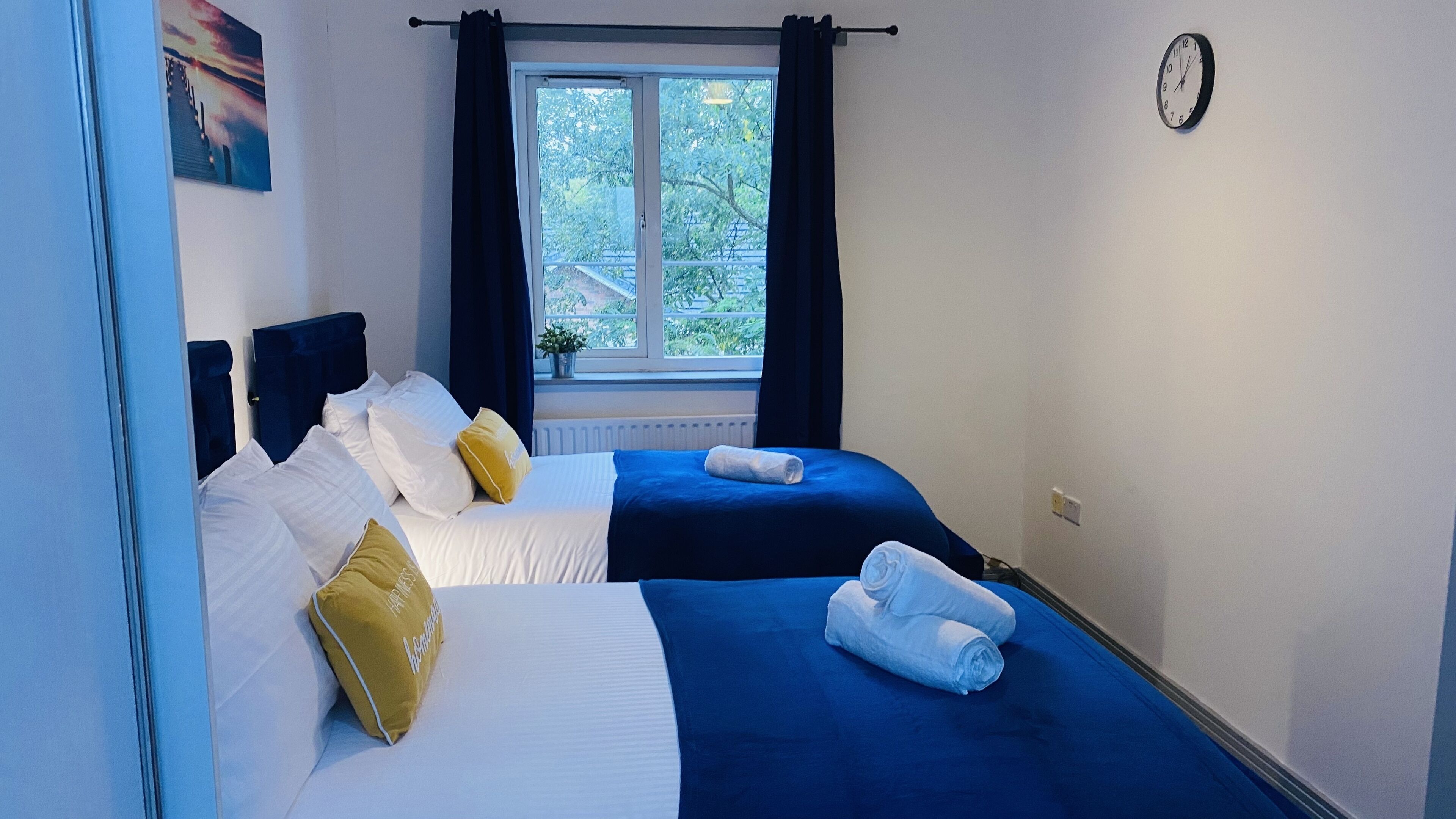 Ein Hotelzimmer mit zwei Betten, einem fenster mit Vorhängen, einer Wanduhr und Blick auf Grün.