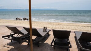 Eine Strandlounge mit Liegestühlen und einem Tisch mit Blick auf das Meer und eine entfernte Küste, wo Reiter zu sehen sind.