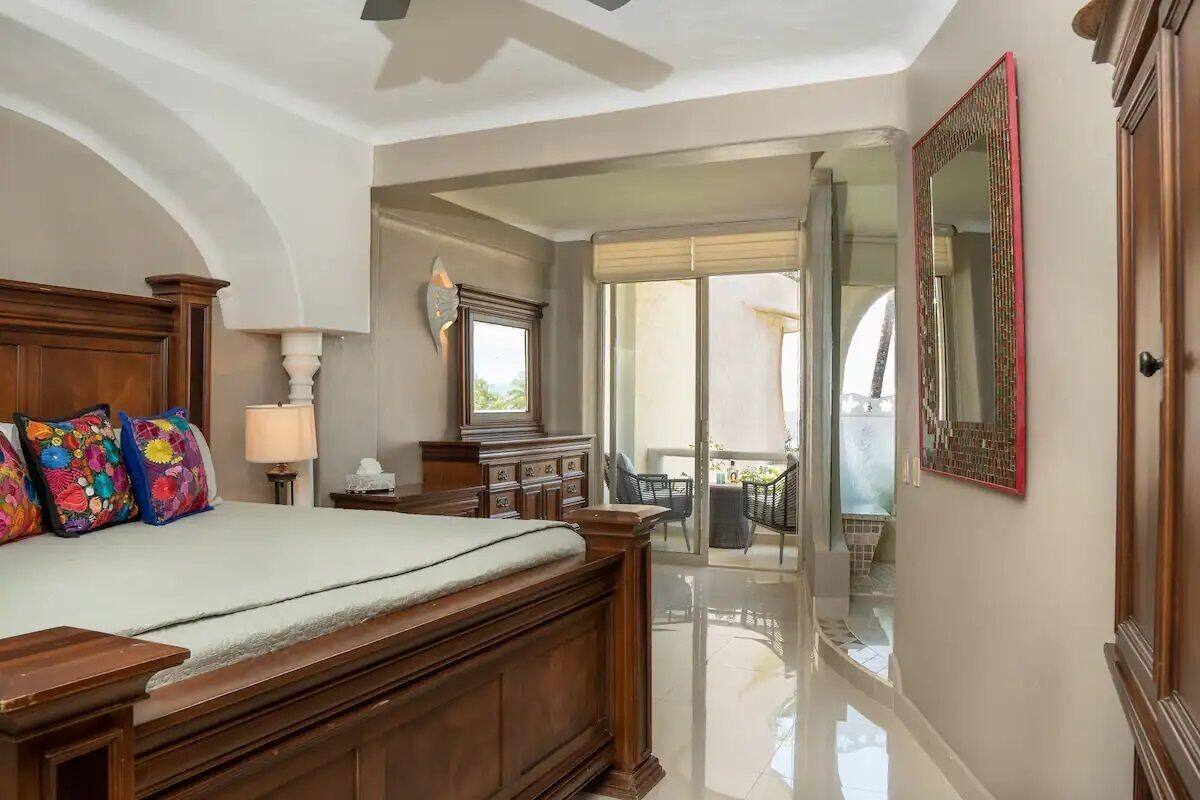 Une chambre avec un grand lit, une table de chevet, une lampe et une commode. Il y a un balcon avec une chaise et une table.