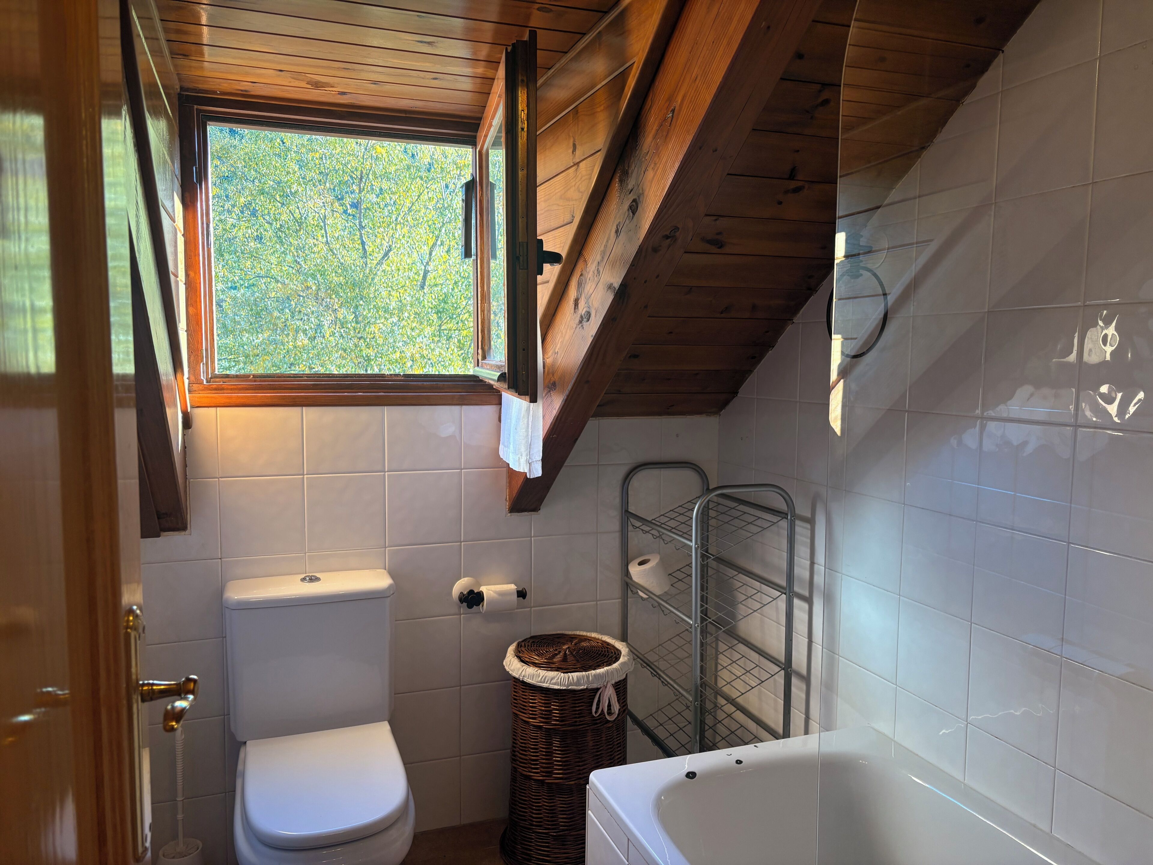 Une salle de bain comprenant des toilettes, une baignoire, un panier à linge en osier et une fenêtre donnant sur les arbres.
