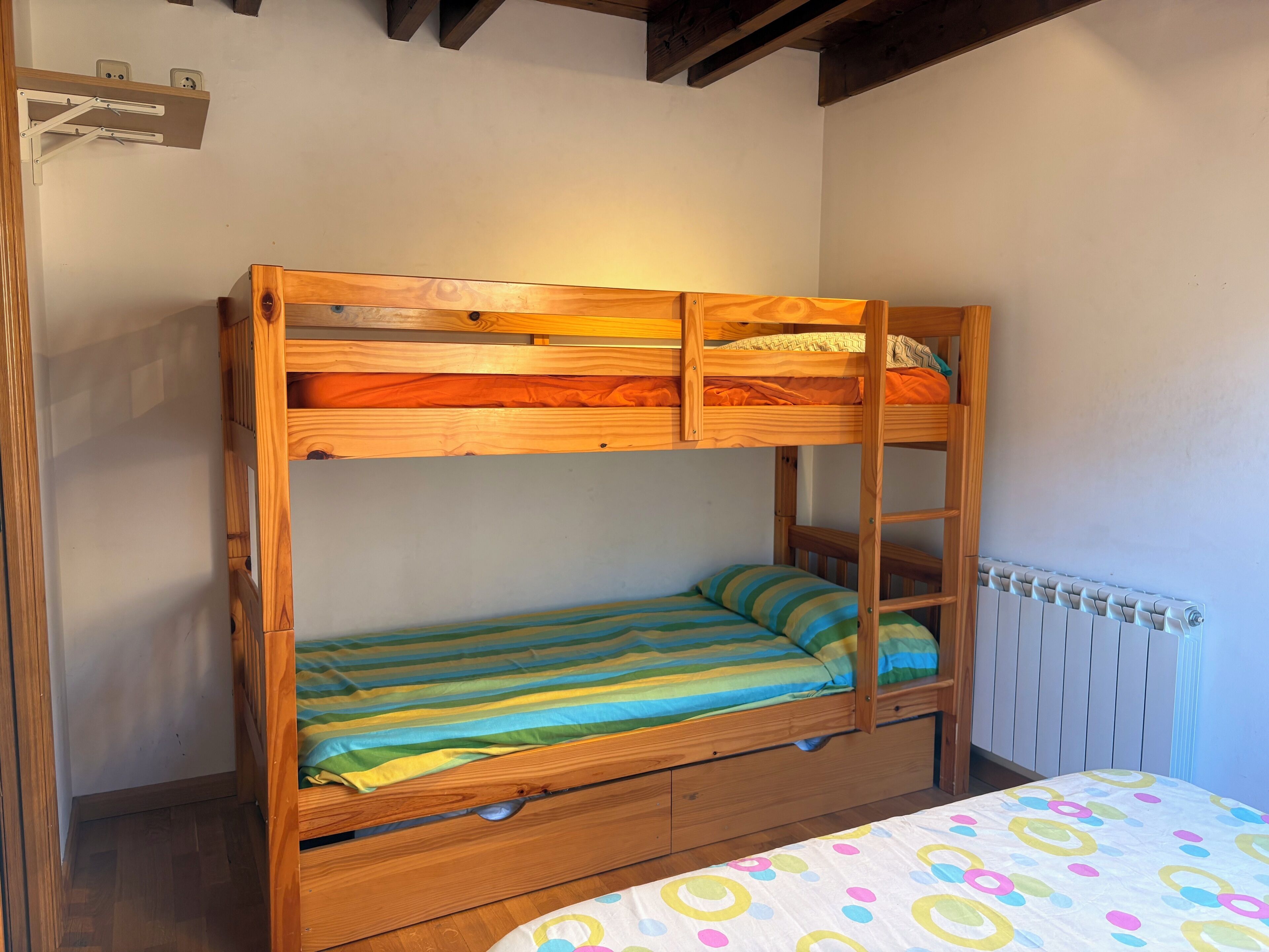 A bunk bed with a lower bunk and a single bed below, wooden walls, a radiator, and a small shelf with electrical outlets.