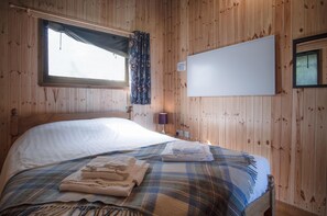 A wooden-paneled room with a bed, a window with curtains, a wall-mounted whiteboard, and a lamp.