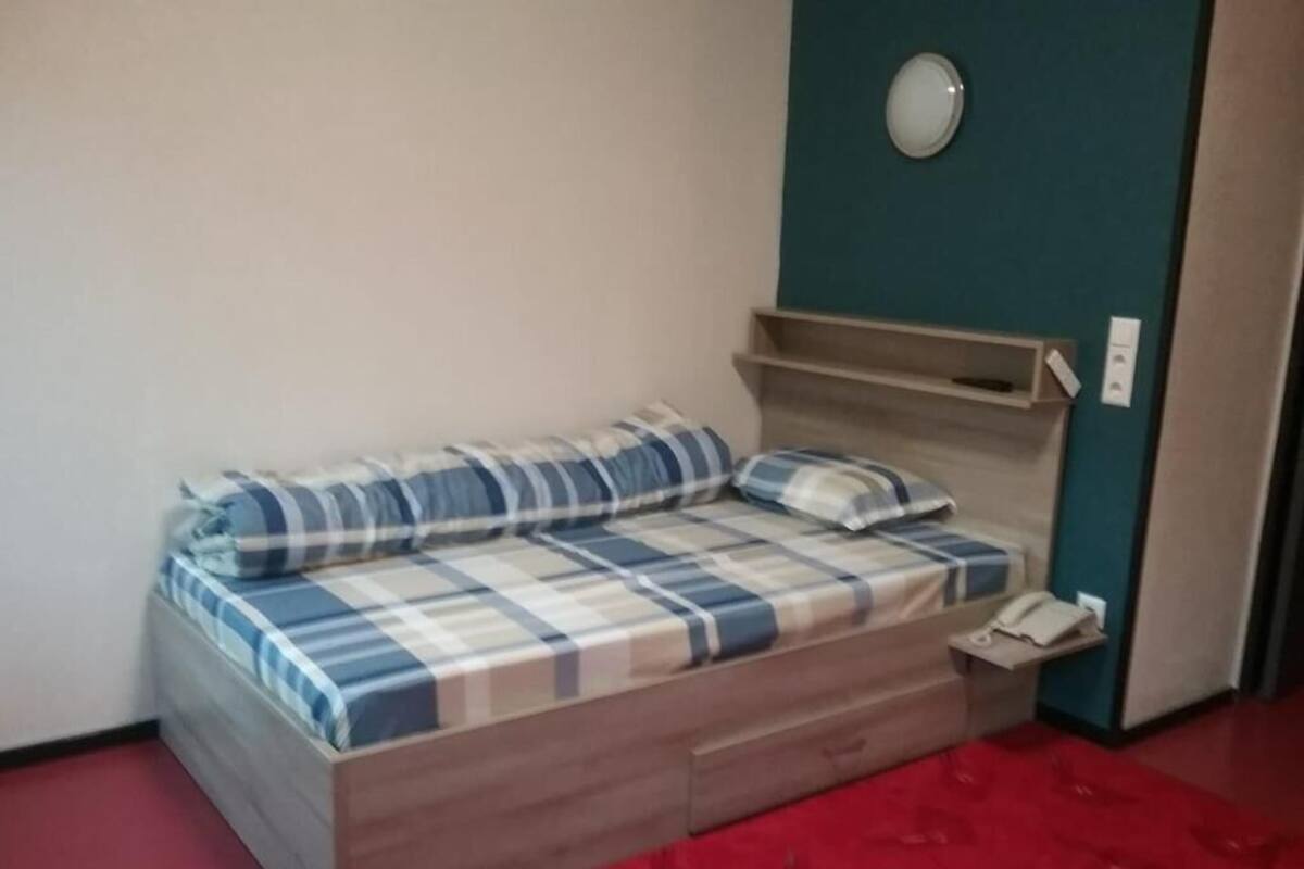 A single bed with a plaid bedspread and pillows, a bedside table with a lamp, and a red carpeted floor.