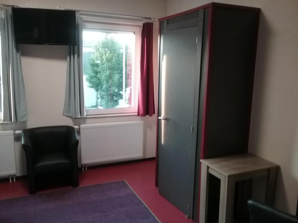 Ein Zimmer mit Fenster, Heizkörper, schwarzem Stuhl, Kleiderschrank und kleinem Tisch.