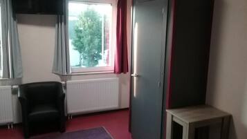 Ein Zimmer mit Fenster, Heizkörper, schwarzem Stuhl, Kleiderschrank und kleinem Tisch.
