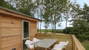 Een houten terras met tafel en stoelen, met uitzicht op een landelijke omgeving met bomen en velden.