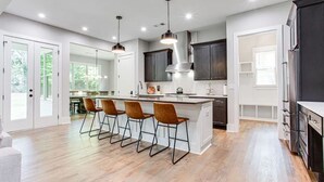 A modern kitchen with a central island, bar stools, pendant lighting, and dark cabinetry. - Beautiful Modern New Home Near Brookhaven Midtown (Atlanta)
