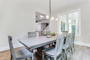 Uma área de refeições com uma grande mesa de madeira e seis cadeiras, um lustre pendurado, e vista para uma cozinha e uma escadaria ao fundo.