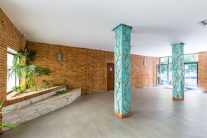 A spacious lobby with a brick wall, two tall columns with intricate designs, a palm tree, and a staircase leading to an upper level.