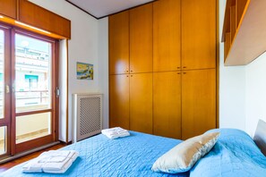 A bedroom with a large wooden wardrobe, a bed with blue bedding, and a window with a view of buildings.