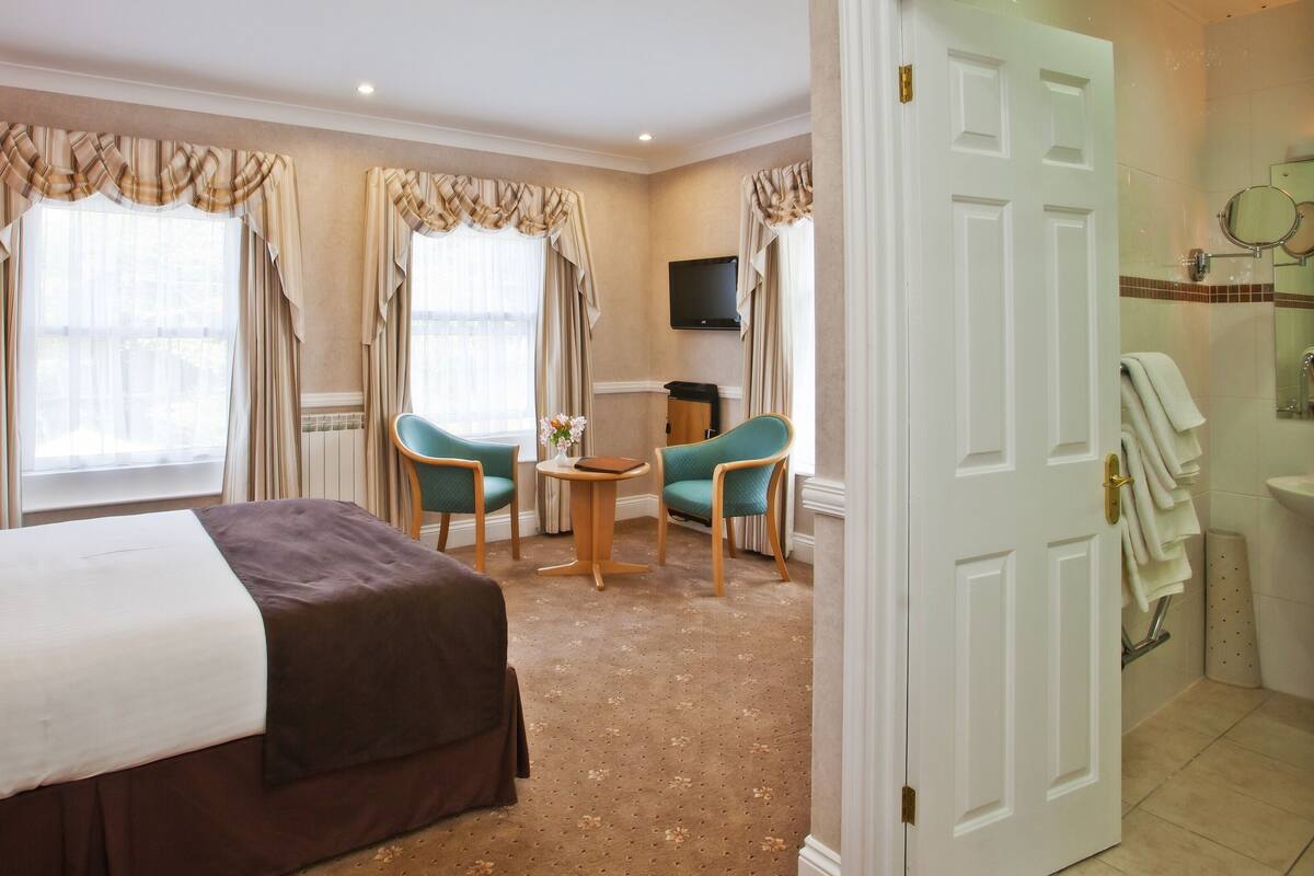 Une chambre d’hôtel comprenant un lit, deux chaises, une petite table, un téléviseur et une salle de bain avec un lavabo et un miroir.