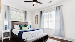 Ein Schlafzimmer mit Bett, Nachttischen, Deckenventilator und großen Fenstern mit Vorhängen.