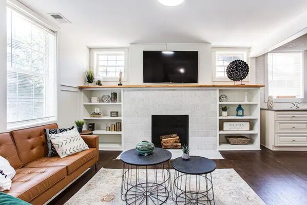 A living room with a fireplace, a TV mounted above it, a brown leather sofa, and two round tables with metal legs.