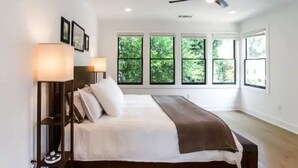 A bedroom with a bed, two bedside lamps, a ceiling fan, and large windows with a view of greenery. - Sleek & Beautiful New Luxury Home In Atlanta (Atlanta)