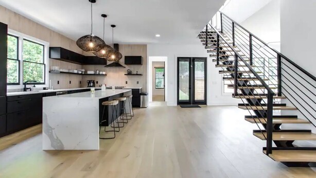 A modern kitchen with a central island, bar stools, pendant lighting, and a staircase with a glass railing. - Sleek & Beautiful New Luxury Home In Atlanta (Atlanta)