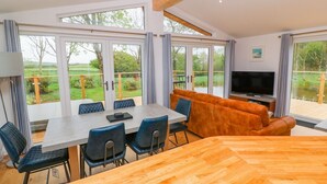 A modern living room with a dining area, large windows, and a TV. - 21 Meadow Retreat (Liskeard)