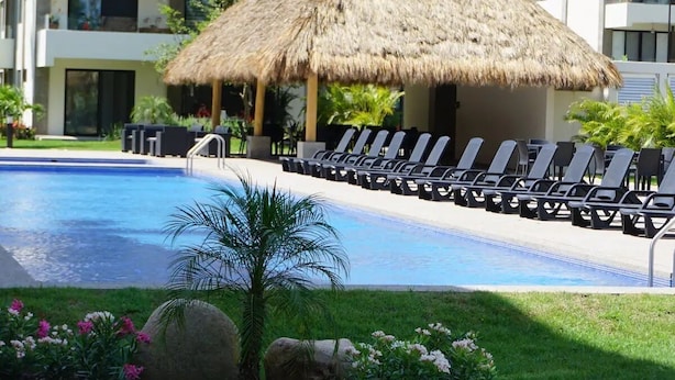 A swimming pool with lounge chairs, a thatched-roof structure, and a modern building in the background. - CONDO LOMA by TRVL2HM (Nuevo Nayarit)