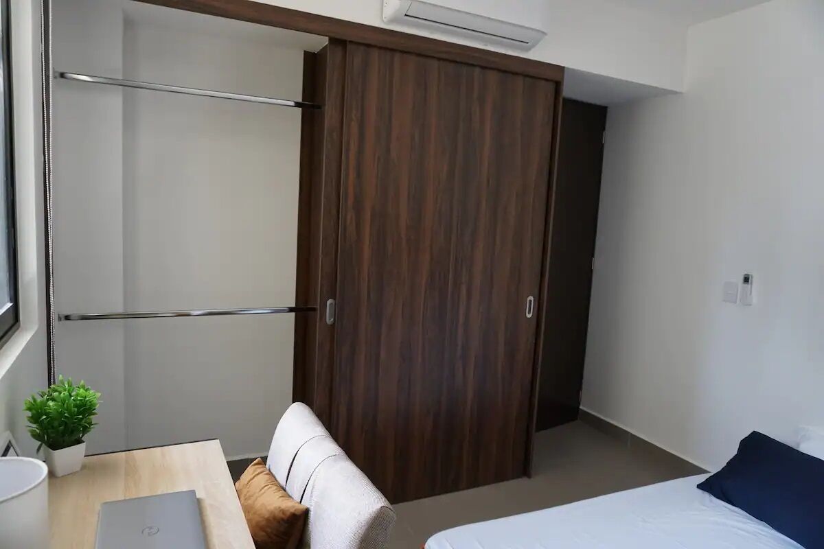Una habitación de hotel moderna con un armario de madera, un escritorio pequeño con una computadora portátil, una cama con almohada azul y una planta en maceta.