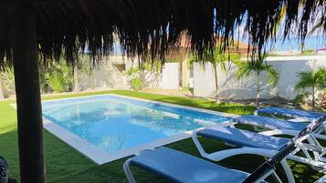 Una piscina con agua azul cristalina, rodeada de césped artificial y sillas reclinables blancas.
