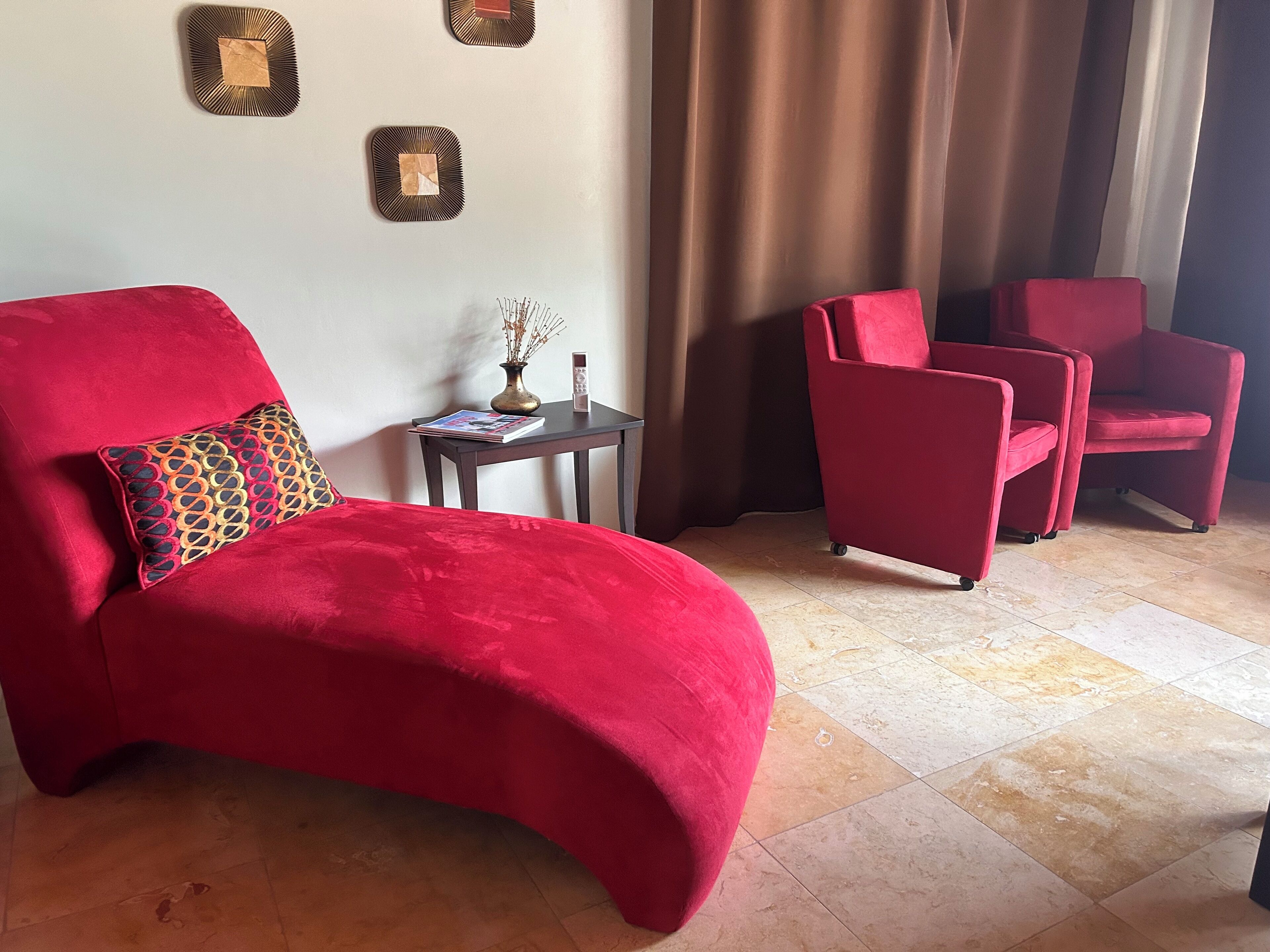 A red armchair with a patterned pillow, a small table with a vase and book, and two red chairs in a room with a tiled floor and a curtain.