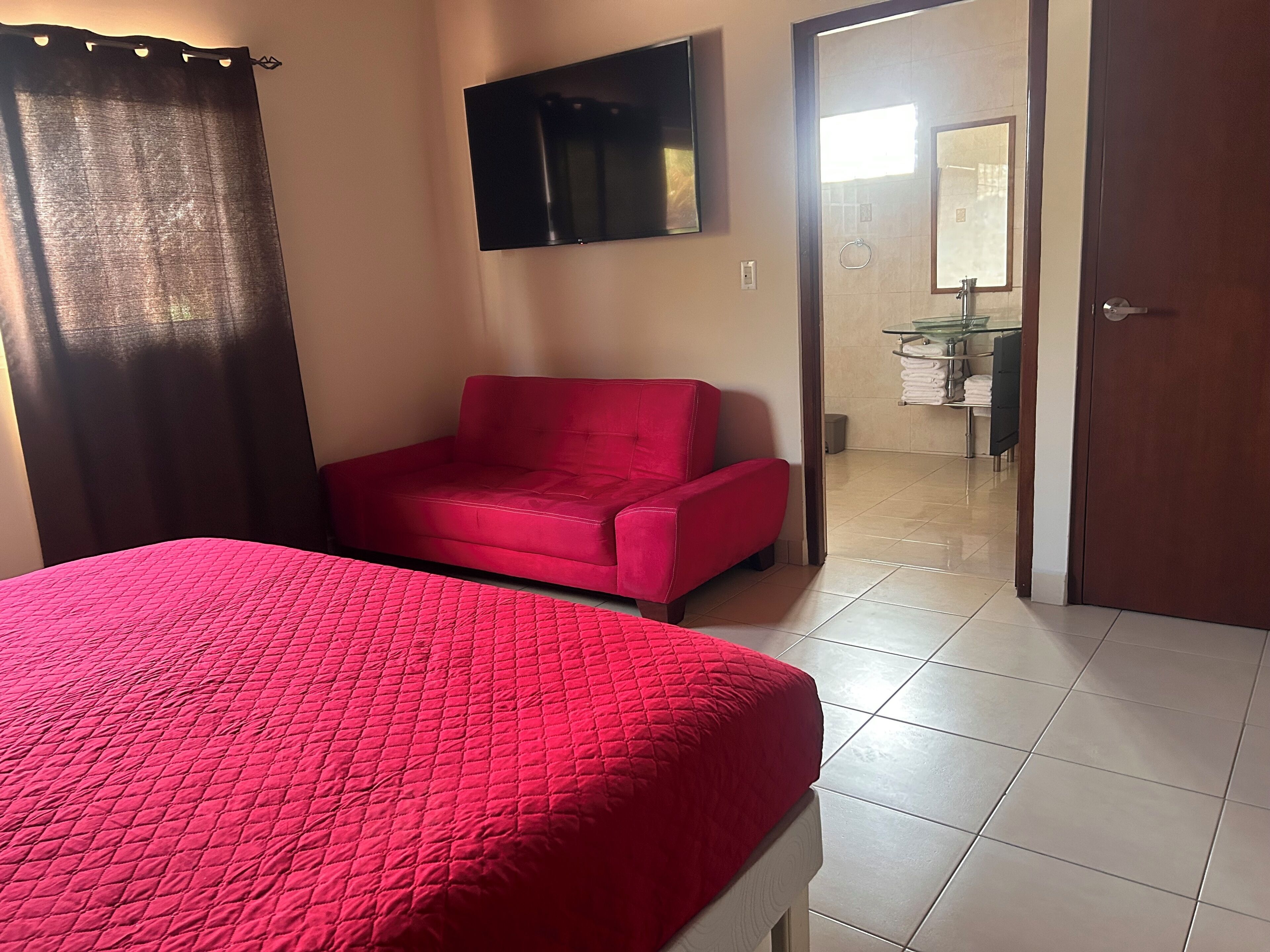 Une pièce comprenant un lit, un fauteuil rouge, un téléviseur fixé au mur et une salle de bain visible à travers une porte ouverte.