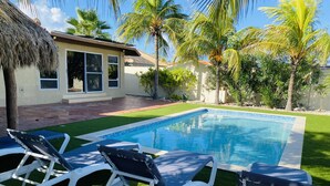 A swimming pool with white lounge chairs, palm trees, and a house with a thatched roof. - Villa Mi Dushi Cas Aruba (Noord)