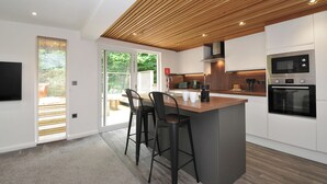Eine moderne Küche mit Holzboden, einer großen Kochinsel mit Hockern, einem Einbauofen und einer Glastür, die zu einem Außenbereich führt.