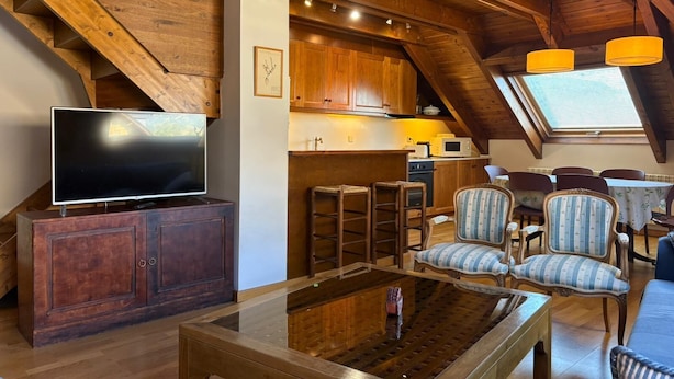 A loft-style room with a television, wooden furniture, a glass coffee table, and a kitchen area with wooden cabinets and a window.