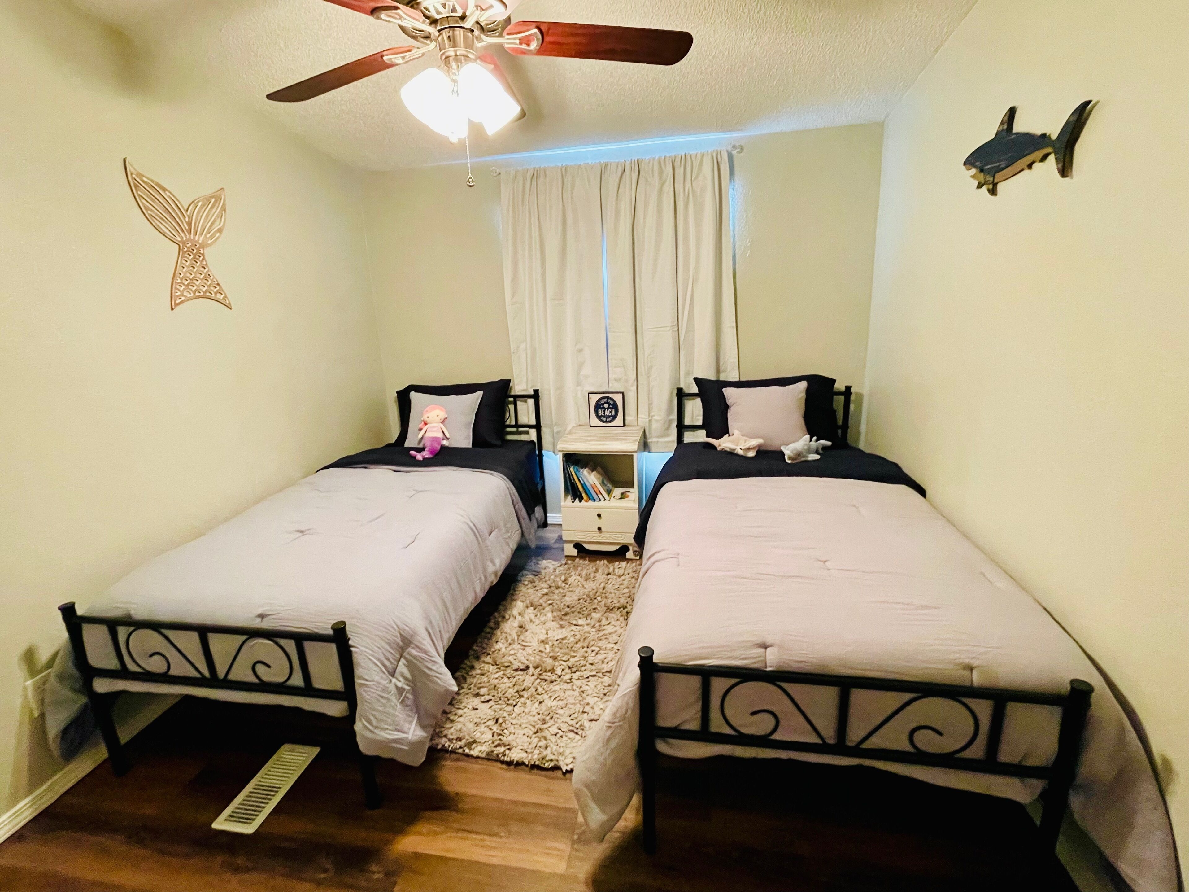 A bedroom with two single beds, a ceiling fan, a nightstand, and a small shelf with books.