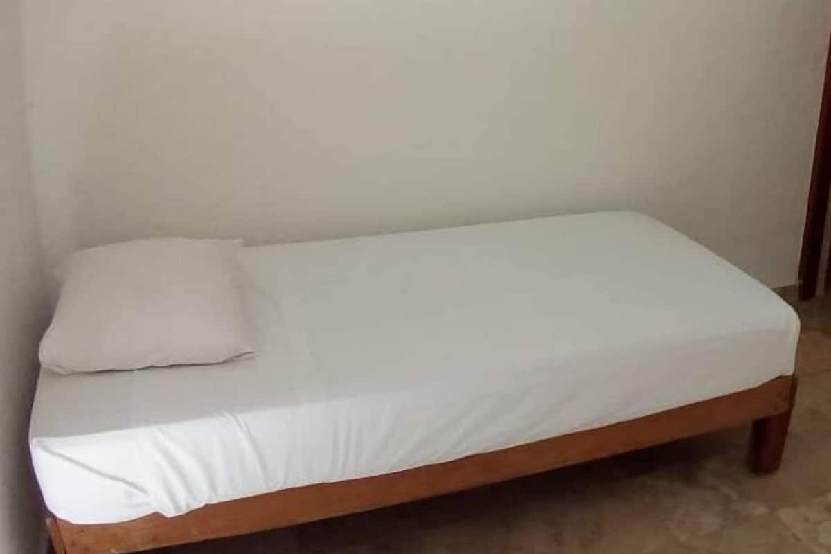 Ein Einzelbett mit Holzbettgestell und weißer Bettwäsche in einem Zimmer mit schlichten Wänden und Fliesenboden.