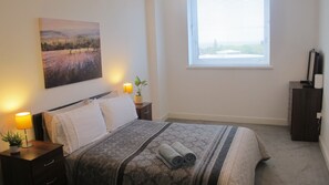 A neatly arranged bedroom with a bed, bedside lamps, a window, and a painting on the wall. - Lovely-cozy Apartment in Brierley Hill (Brierley Hill)