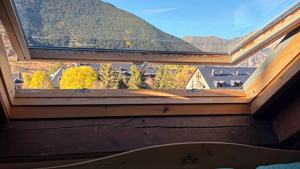 Une pièce dotée d’une lucarne, d’un plafond en bois et offrant une vue sur une montagne et des bâtiments.