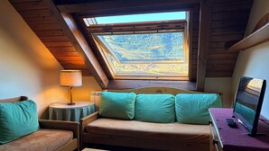 Une pièce chaleureuse avec un plafond en pente, un canapé agrémenté de coussins turquoise, une petite table, une lampe et un ordinateur posé sur un bureau.