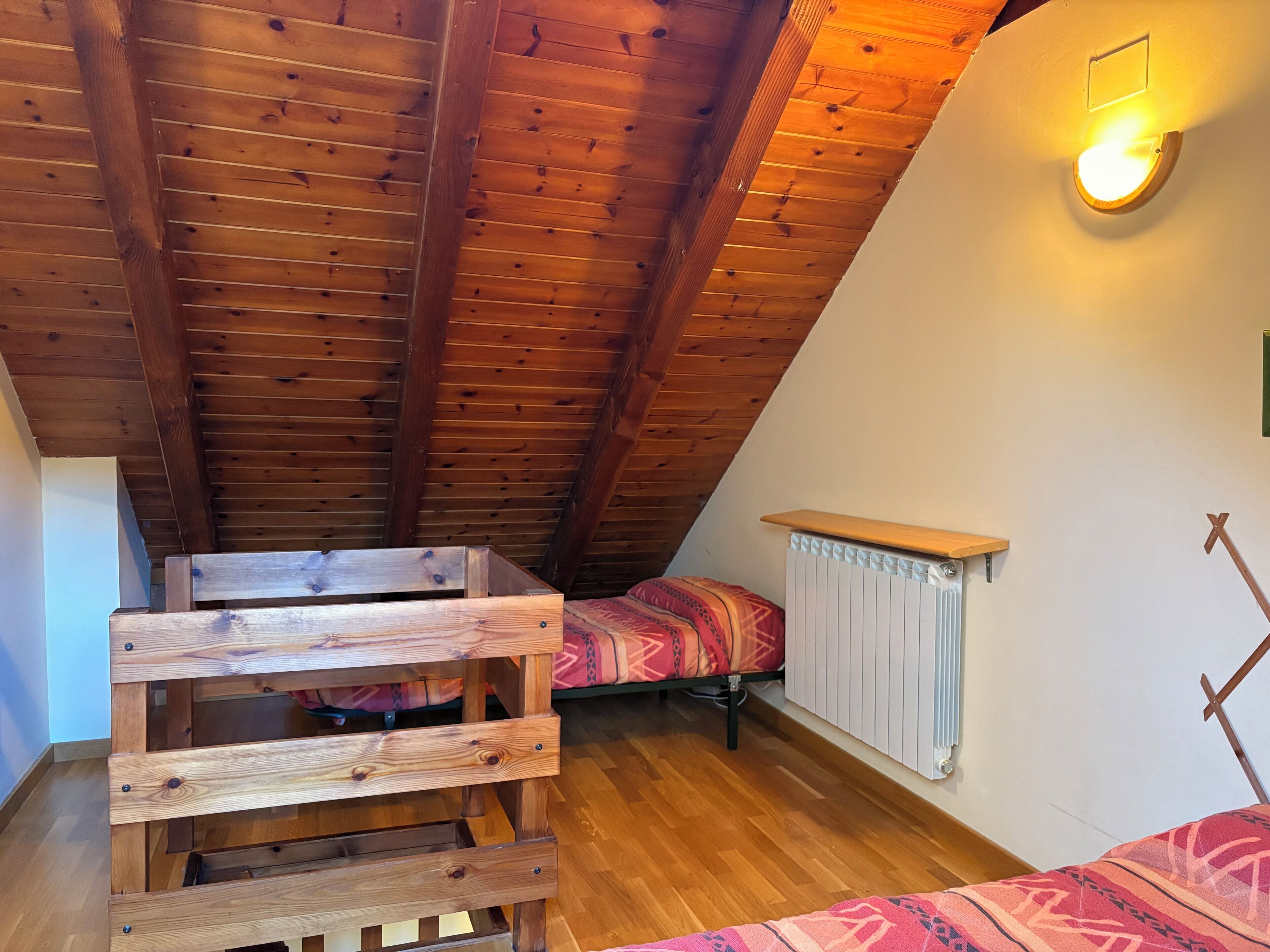 Une chambre à coucher aménagée dans un espace en hauteur, avec un plafond en bois, un lit recouvert d’une literie à motifs, un banc en bois et un luminaire fixé au mur.