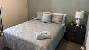 A neatly made bed with a patterned bedspread and pillows, a bedside lamp, and a small potted plant on a nightstand.