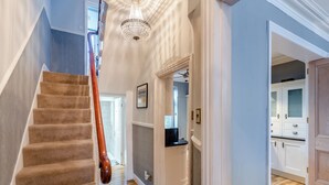 A staircase with a wooden handrail leading to a room with a chandelier, wooden floors, and a kitchen area with white cabinets.