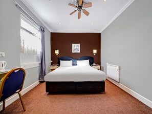 Une chambre d’hôtel avec un grand lit, un bureau avec une chaise, un ventilateur de plafond et une fenêtre avec des rideaux.