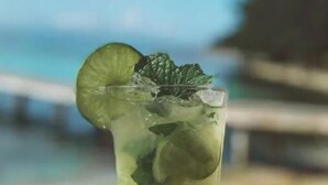Un grand verre rempli d’une boisson verte, garni d’une tranche de citron vert et de quelques feuilles de menthe, posé sur une table en bois, près de la plage.